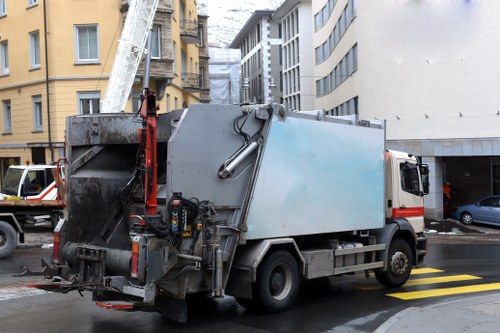 A large commercial waste collection truck parked on a city street in Mill Hill, featuring a grey and blue metal body designed for rubbish collection. The truck has a hydraulic lift at the rear for lifting and emptying waste bins, with visible hoses and mechanical components along the sides. The vehicle is situated next to a pedestrian crossing with yellow stripes on the pavement, and nearby modern multi-storey buildings, some with balconies, are seen in the background. On the right side of the image, parked cars are visible, and a construction or scaffolding structure with a protective covering is attached to a building in the background. The scene is captured during daylight hours, with overcast lighting creating soft shadows, reflecting typical urban waste management activity in Mill Hill. This image is relevant to commercial waste removal services provided by companies such as Commercial Waste Removal Mill Hill, focusing on rubbish collection in the local area.