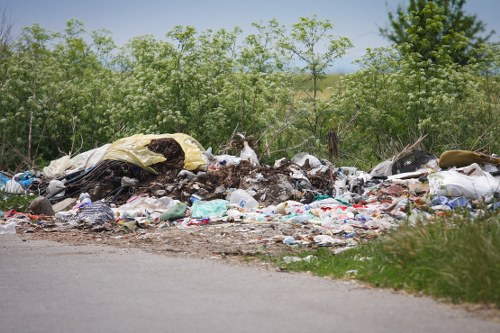 A large roadside pile of mixed waste and rubbish, including discarded plastic bags, paper, and fabric materials, situated next to a grassy verge with dense green shrubbery and small trees in the background. The waste appears to be spilled onto the ground and partially extends onto the edge of a paved road, with some items visibly weathered and partially decomposed. The scene suggests illegal dumping or improper waste disposal in an outdoor environment near Mill Hill, illustrating the type of rubbish that commercial waste removal services like those offered by Commercial Waste Removal Mill Hill aim to manage and clear efficiently.