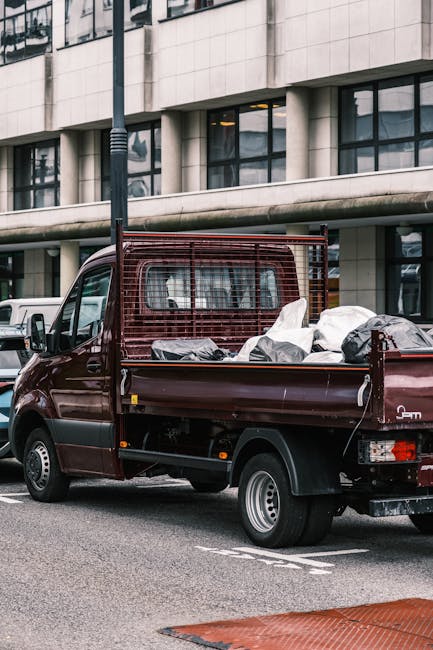 A small burgundy flatbed truck parked on a city street in front of a modern building with large glass windows and concrete exterior walls. The truck's open cargo area is filled with various types of w