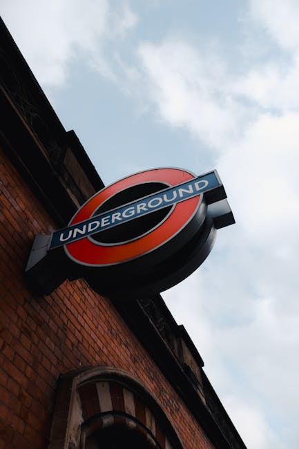 A close-up photograph of a circular London Underground sign mounted on the exterior brick wall of a building. The sign features a prominent red circle with a horizontal blue bar across the middle, whi